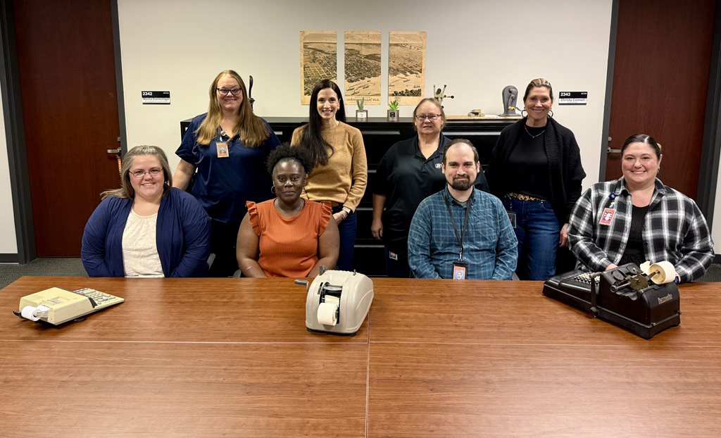 Photo of the team members of the Clerk's finance team sitting and standing behind a wooden table with calculators from various eras sitting in front of them