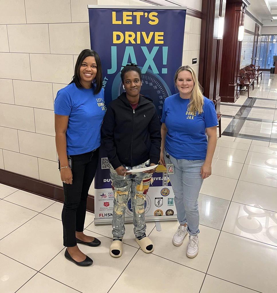 Let's Drive Jax program participant with Clerk employees in front of Let's Drive Jax sign